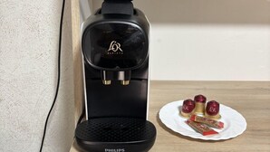 A black coffee maker on a wooden shelf with a white plate of food and a remote control.
