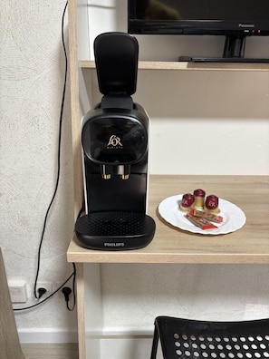 A black coffee maker on a wooden shelf with a white plate of food and a remote control. - The Secret Nest (Arras)