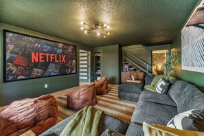 A living room with a large flat-screen TV displaying a Netflix interface, a sofa, leather poufs, and a patterned rug.