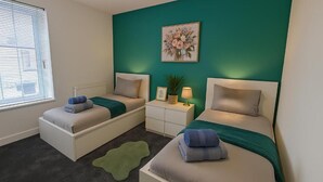A bedroom with two single beds, a nightstand with a lamp, a framed picture, and a green rug. - Armstrong St - Cosy 3 Bedroom Home (Workington)