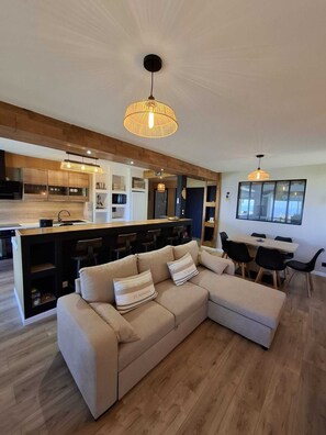 Eine moderne Küche mit Kochinsel, ein Essbereich mit Tisch und Stühlen sowie ein großes L-förmiges Sofa mit Kissen.