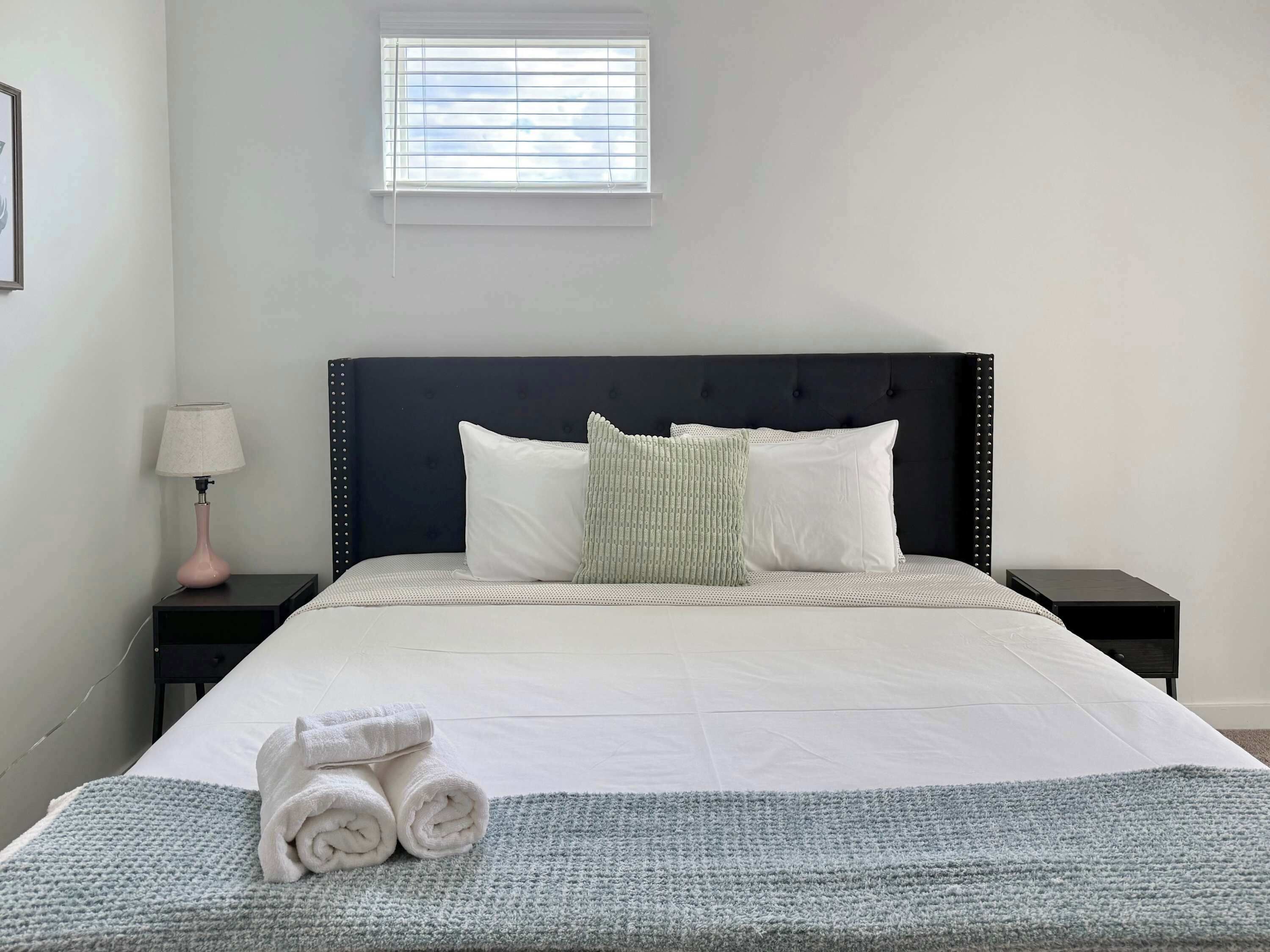A neatly made bed with pillows, a bedside lamp, and a window with blinds.