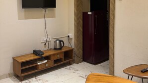A room with a flat-screen TV mounted on the wall, a wooden console table with a kettle, a refrigerator, and a round wooden table with a remote control. - Vijaya's Homestay Indore (Indore)