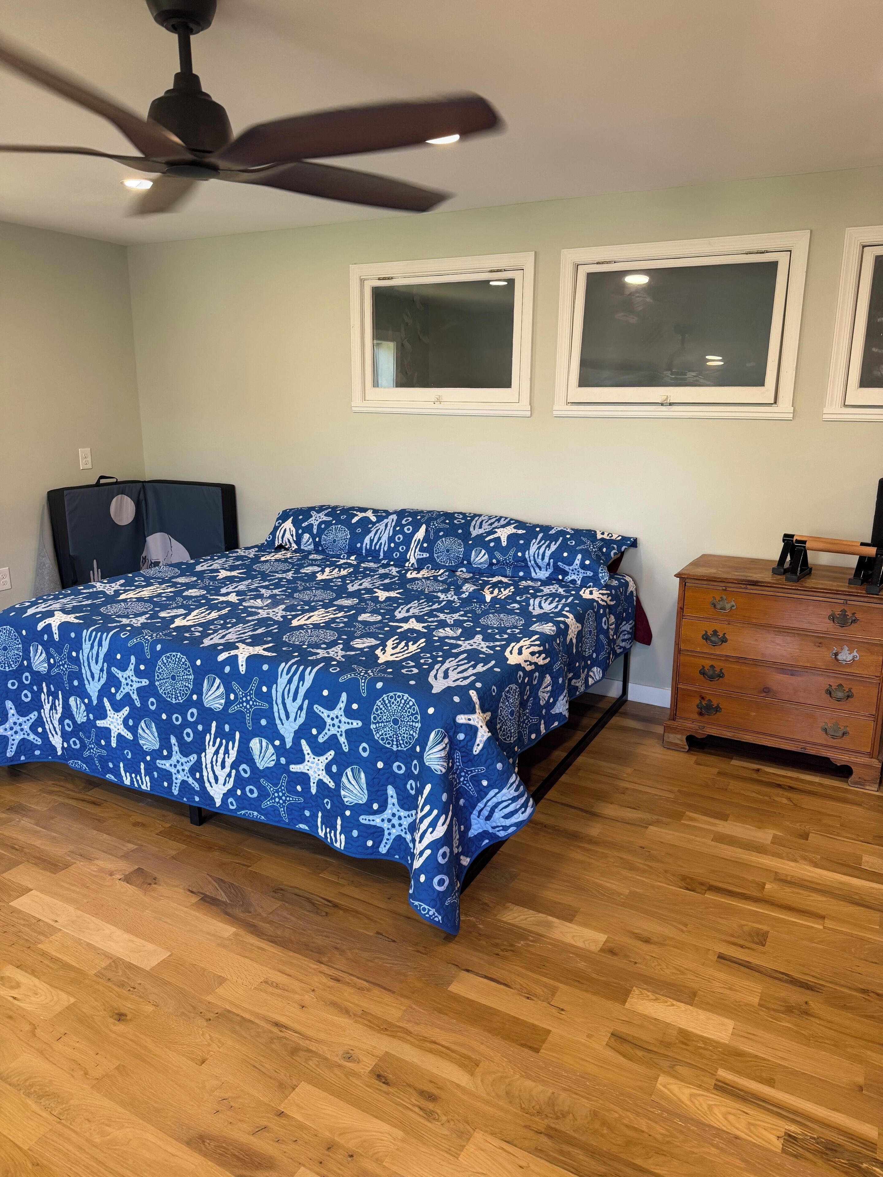 Un dormitorio con cama, cómoda, ventilador de techo y dos ventanas.