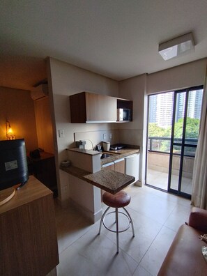 A compact kitchen with a microwave, a sink, and a small countertop. There is a wooden cabinet above the counter and a television on a wooden stand. A stool is placed in front of the counter, and a sliding glass door leads to a balcony. - Studio Urbano-flat confortável-localização nobre (Goiânia)