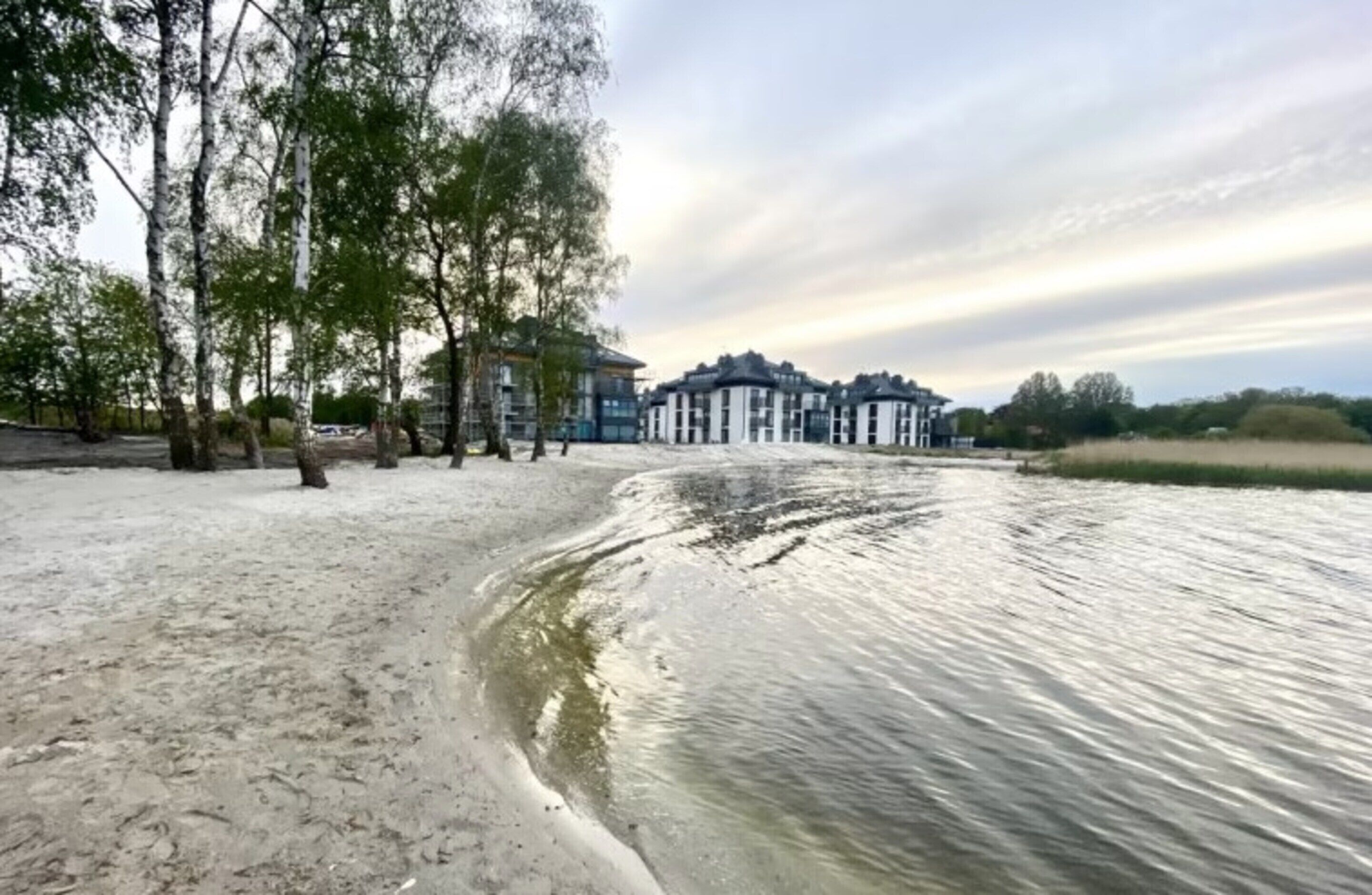 A riverside view with a sandy beach, trees, and a row of buildings in the background.