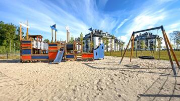 A playground with slides and swings on a sandy area, surrounded by buildings.