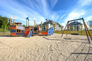 A playground with slides and swings on a sandy area, surrounded by buildings. - Waterview Retreat for 2 in Dziwnow (Dziwnów)