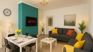 Una sala de estar moderna con comedor, televisor montado en una pared verde aguamarina, y sofá con cojines amarillos y naranjas.