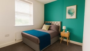 Un dormitorio con cama, una mesita de noche con lámpara, ventana con persianas y una pared de acento en color verde azulado.