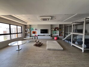 Ein modernes Wohnzimmer mit Fernseher, Regalen mit Spielzeug, einer Kinderecke und einem großen Fenster.