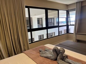 Ein Hotelzimmer mit großem Fenster, einem Bett, einem Nachttisch und Blick auf das gegenüberliegende Gebäude.
