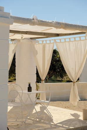 A white metal table with a bottle, a chair, and a white canopy structure. - Trullo Moi (Ostuni)