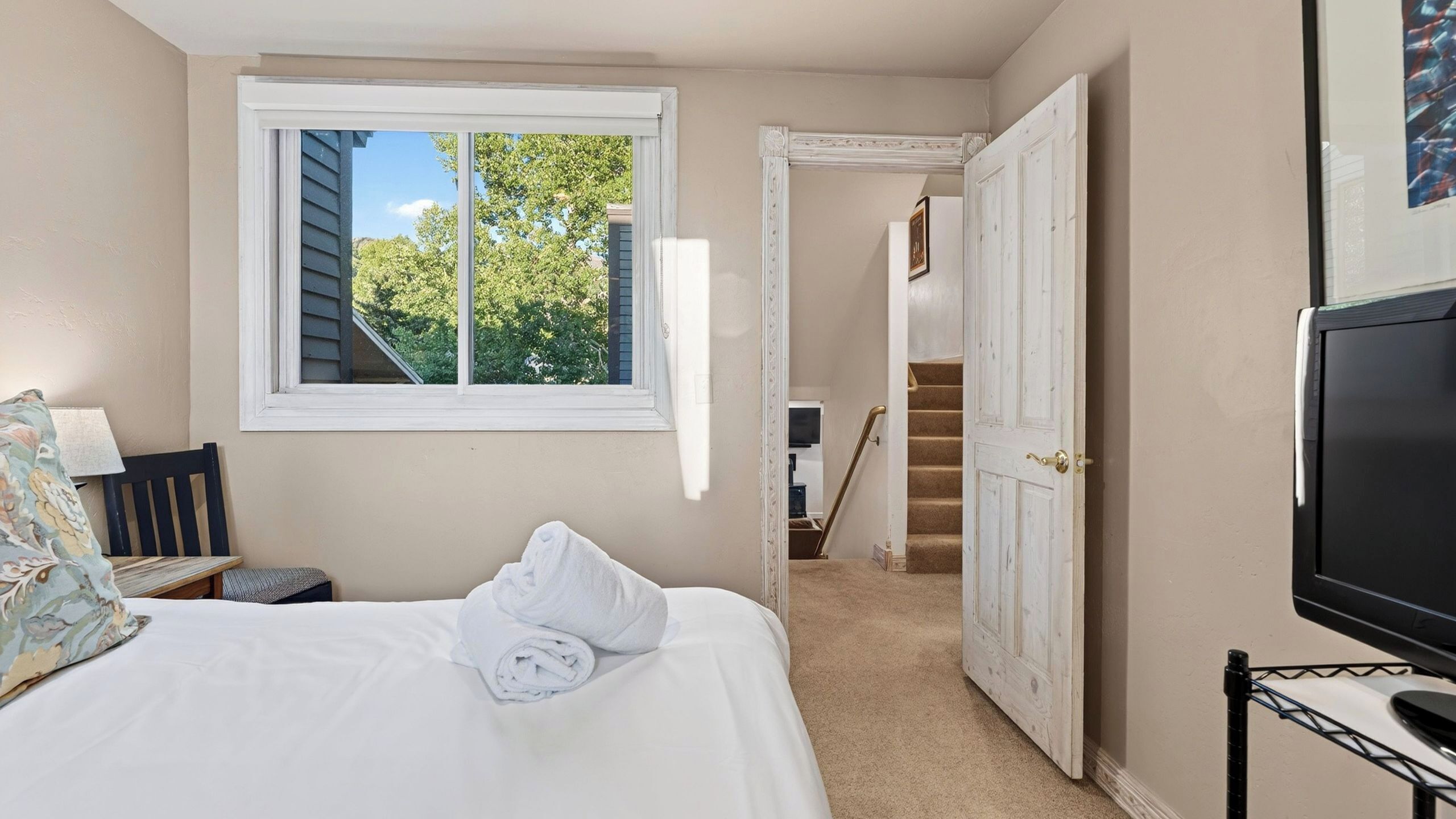 Un dormitorio con cama, ventana, puerta que da a una escalera y televisor sobre un mueble.
