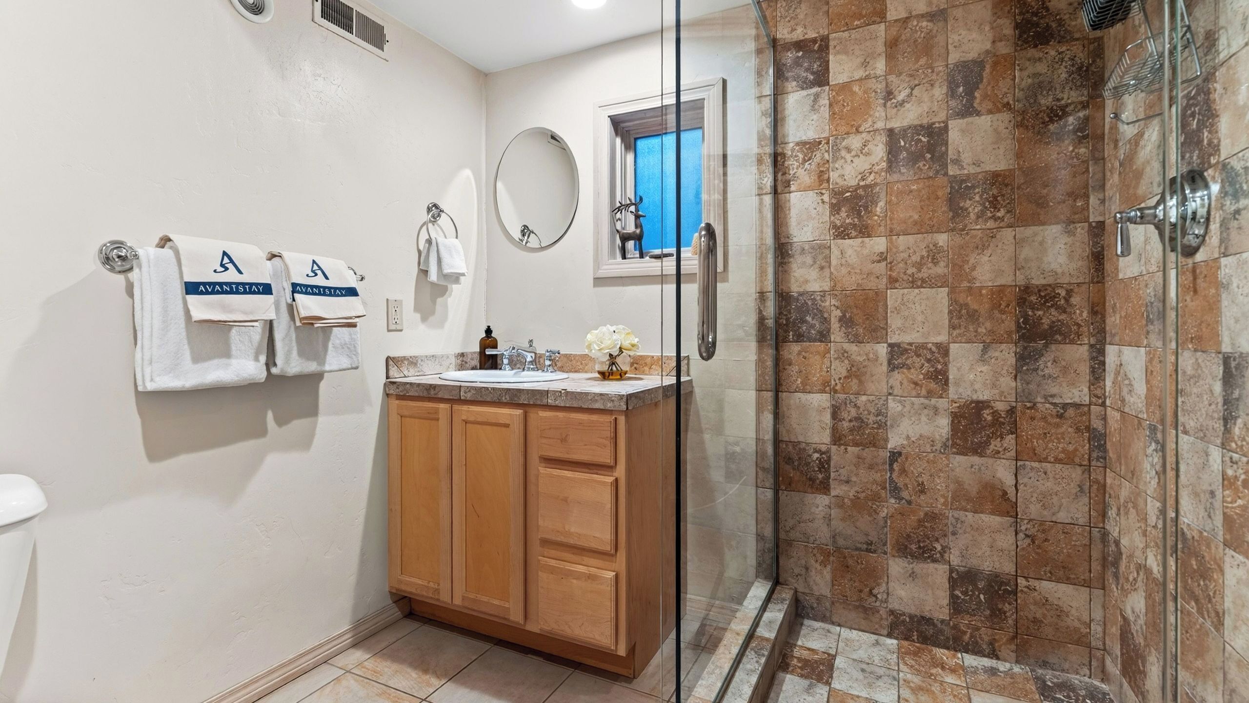 A bathroom with a glass-enclosed shower, a sink with a marble countertop, wooden cabinets, and two white towels with 'AVANTSTAY' branding.