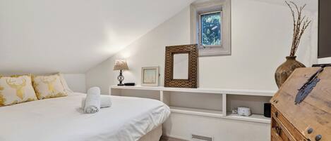 Un dormitorio con una cama, una mesita de noche con lámpara, un marco de cuadro, un ventanal pequeño y un aparador de madera.