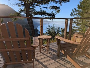 A wooden deck with Adirondack chairs and a small table, overlooking a body of water and mountains. - Blue whale cove | Cliffside Ocean Views | Hot tub (Whitethorn)