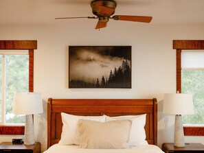 A bedroom with a wooden headboard, a ceiling fan, a framed picture of a forest, and two bedside lamps. - Blue whale cove | Cliffside Ocean Views | Hot tub (Whitethorn)