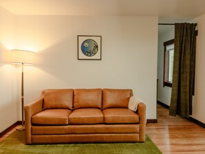 Een kamer met een bruine leren bank, een vloerlamp, een ingelijste foto aan de muur en een raam met gordijnen.