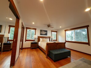 A bedroom with a large bed, bedside tables, a nightstand, a bench, a ceiling fan, and windows with a view of trees. - Blue whale cove | Cliffside Ocean Views | Hot tub (Whitethorn)