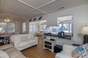 Ein geräumiges Wohnzimmer mit Flachbildfernseher, weißen Möbeln und Blick auf den Strand durch das Fenster.
