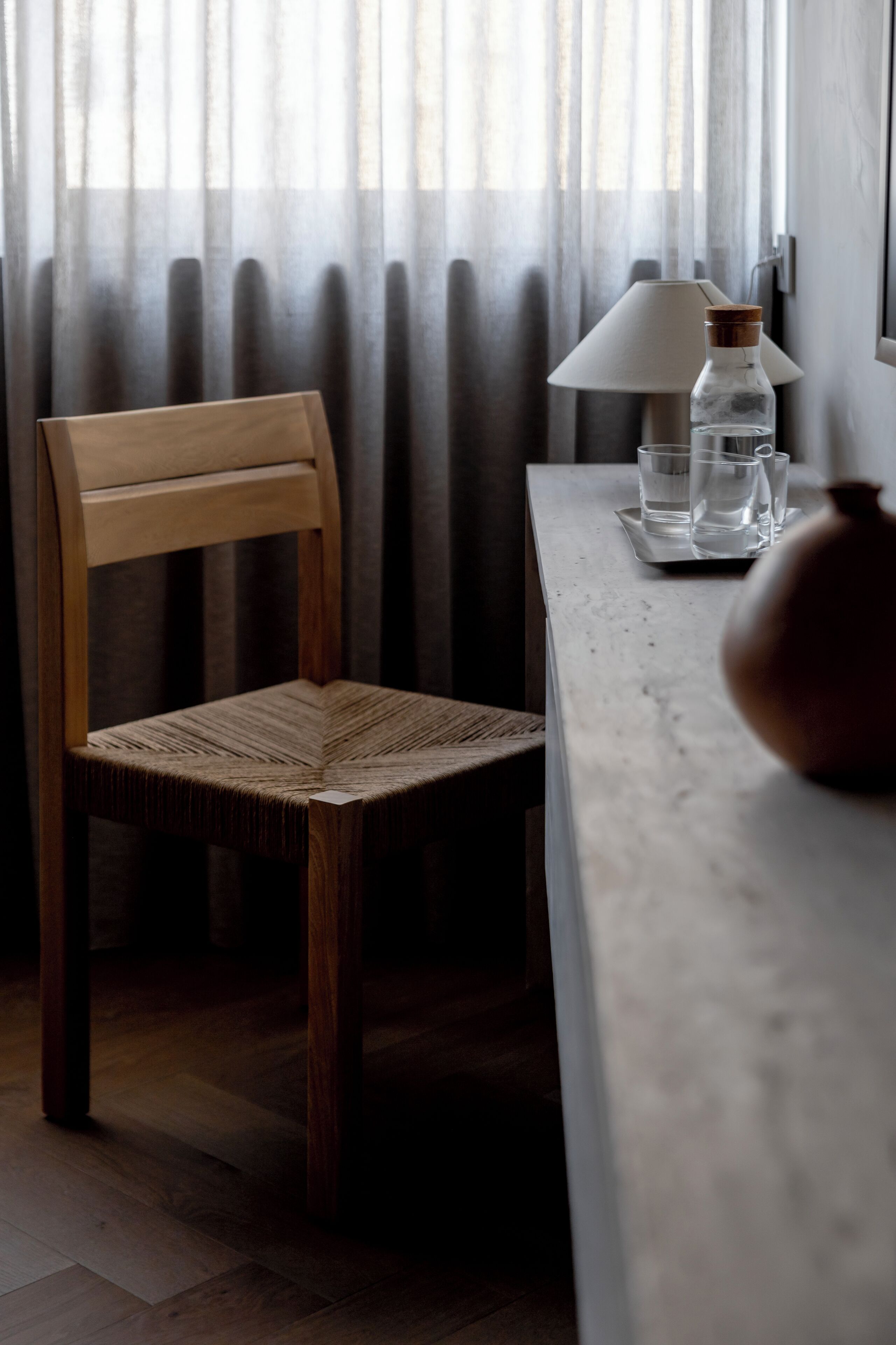 A wooden chair with a woven seat, a table with a lamp, a bottle, and a vase.