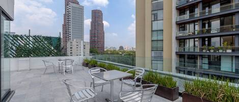 A rooftop terrace with outdoor furniture, a glass balustrade, and a view of surrounding buildings.