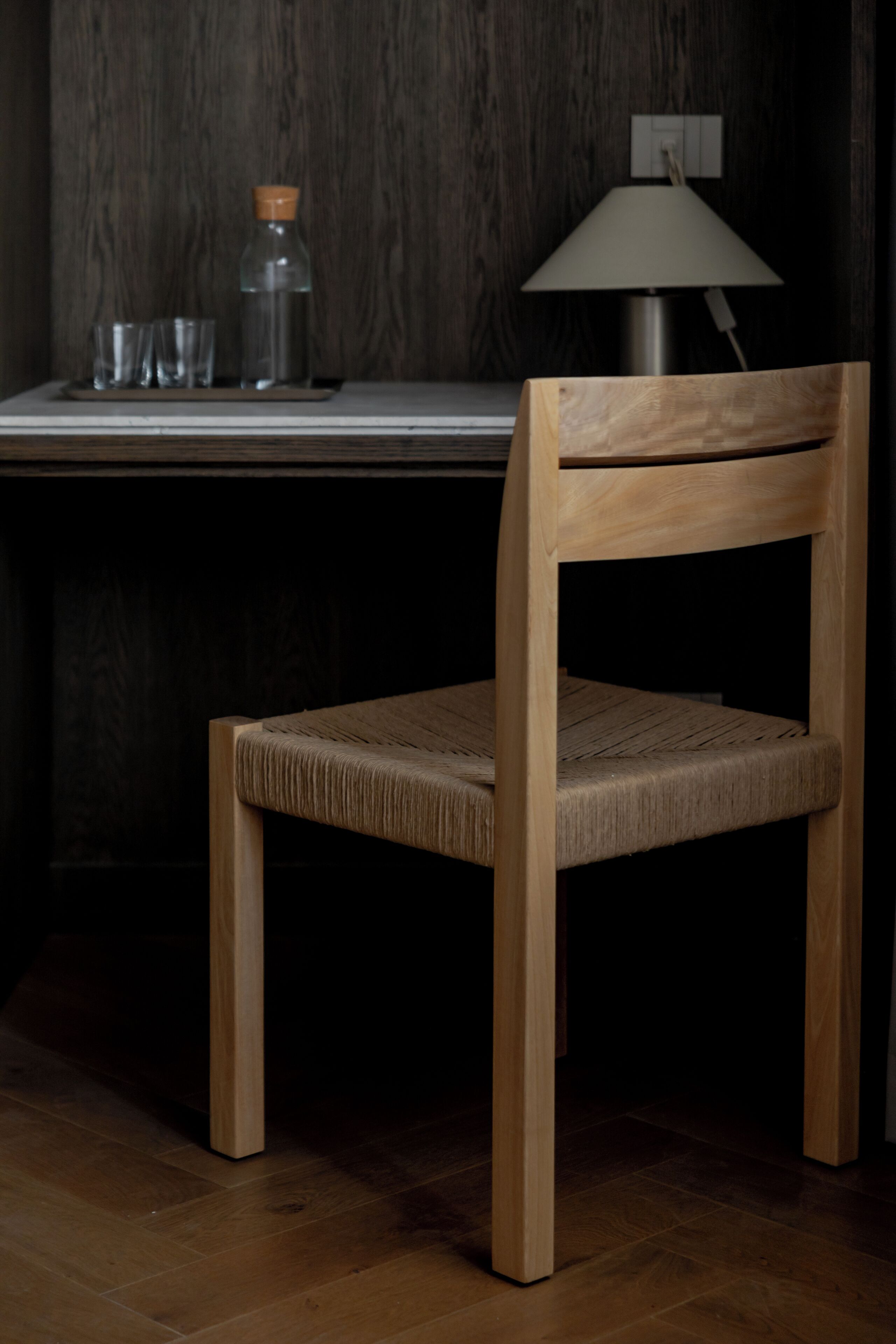 A wooden chair with a woven seat and backrest, positioned in front of a desk with a lamp and a bottle.