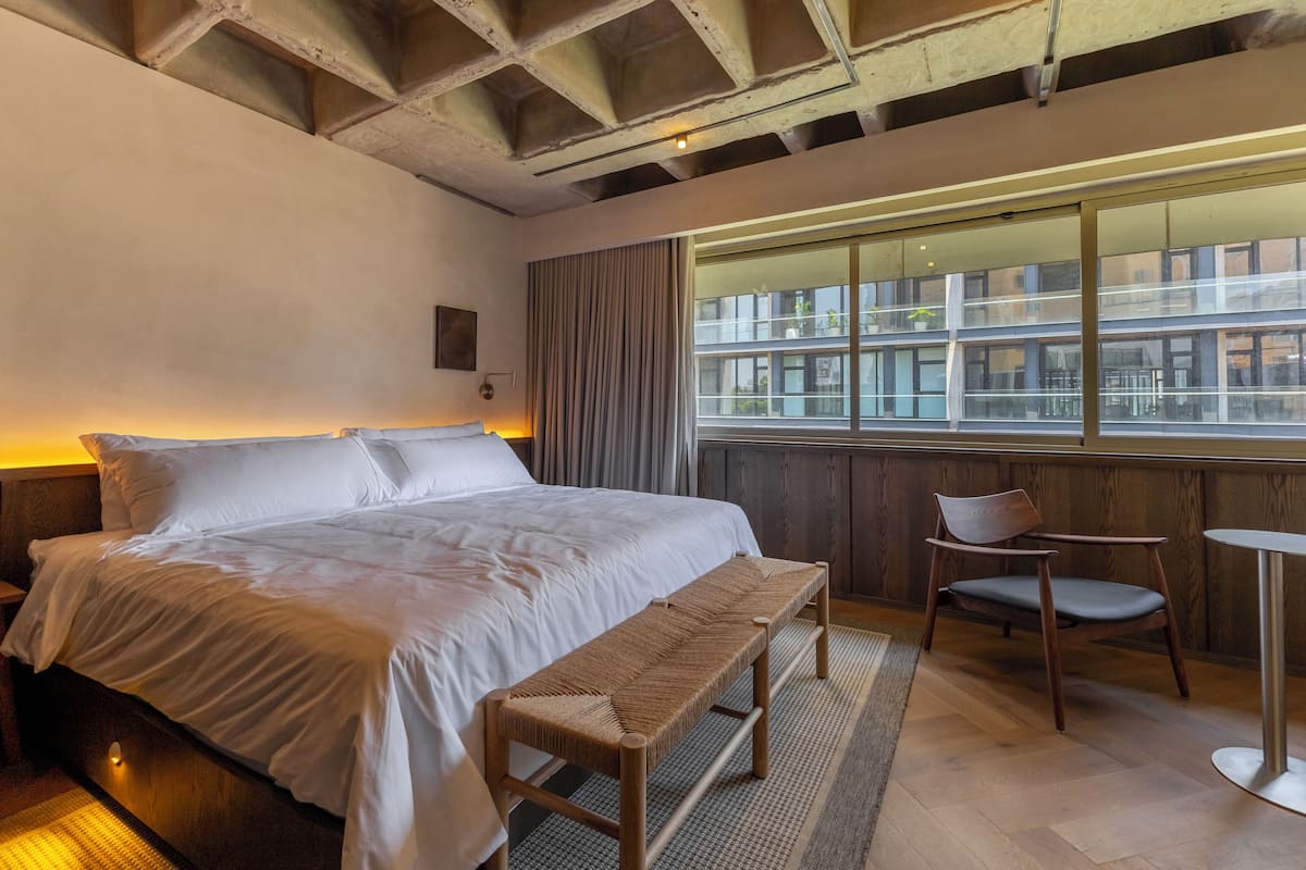 Une chambre d’hôtel avec un grand lit, une banquette, une chaise, une petite table et une fenêtre donnant sur des immeubles.