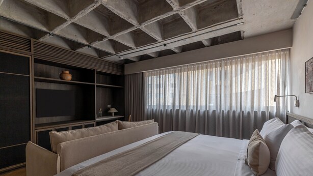 Une chambre à coucher moderne avec un grand lit, une étagère en bois ornée d’un vase et une fenêtre avec des rideaux légers.