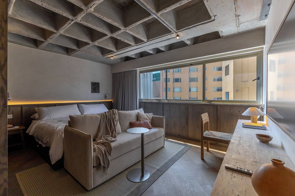 Une chambre d’hôtel moderne, équipée d’un lit, d’un canapé, d’une petite table et offrant une vue sur le paysage urbain grâce à de grandes fenêtres.