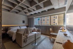 Une chambre d’hôtel moderne, équipée d’un lit, d’un canapé, d’une petite table ronde et offrant une vue sur la ville grâce à de grandes fenêtres.