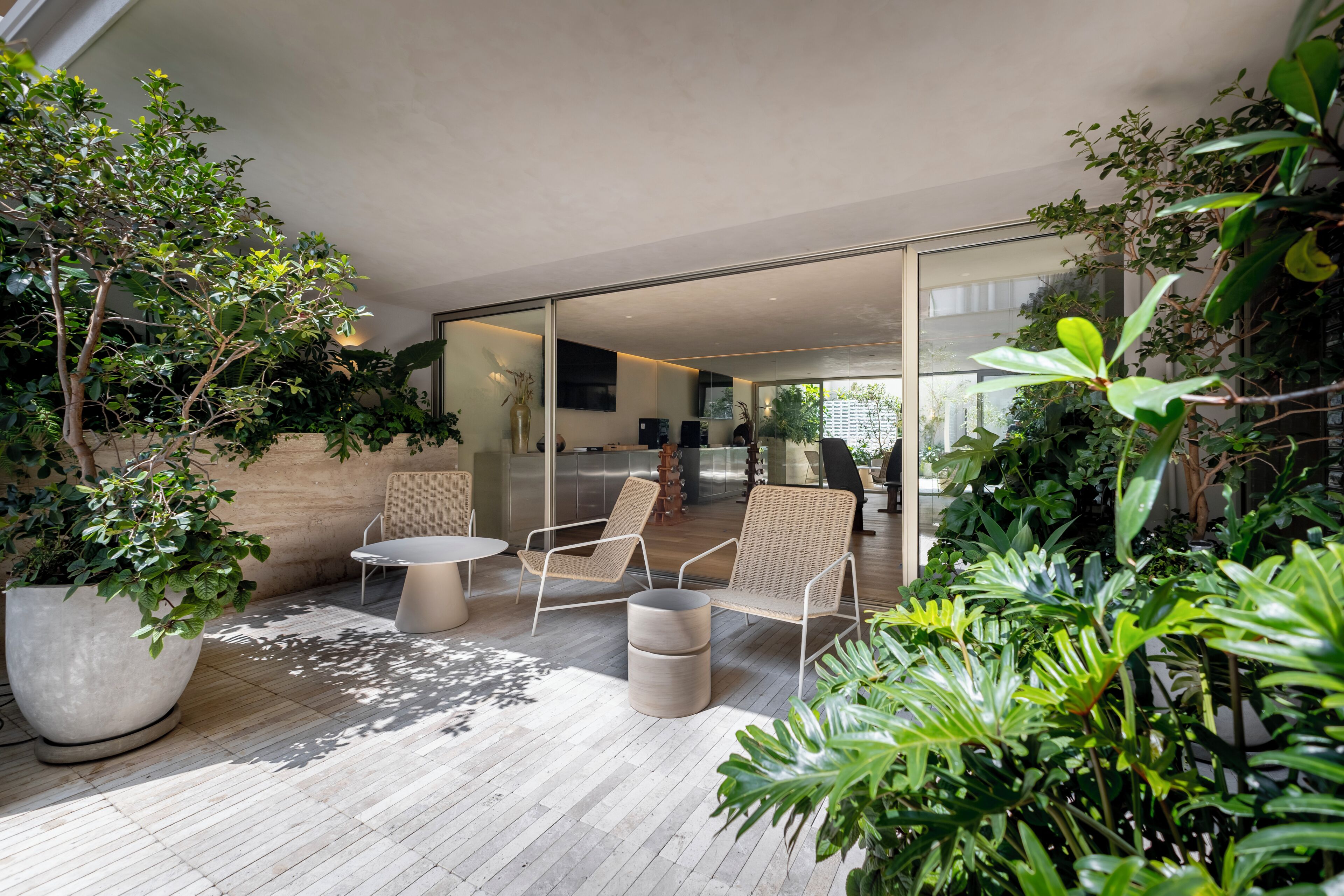 An outdoor patio with a round table and two wicker chairs, surrounded by potted plants and a glass door leading to an interior space.