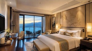 A hotel room with a large bed, a desk with a tray of fruits, a chair, and a view of the sea.