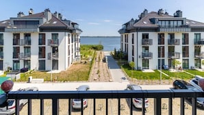 A residential building with a view of a body of water, featuring balconies, a paved pathway, and parked cars. - Premium Dziwnow Coastal Apartment for 3 (Dziwnów)