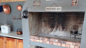 A rustic outdoor bar with a brick fireplace, copper pots, and a sign reading 'BACKYARD BAR.'