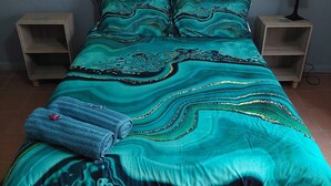 A bed with a teal patterned bedspread, two pillows, and a folded towel. There are two bedside tables with lamps.