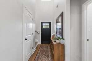 A hallway with a wooden floor, white walls, a black door, and a small wooden console table with a plant and candles. - Gathering Home - Sleeps 10 - 4 King Beds 4.5 Baths (College Station)