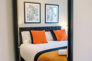Een net opgemaakt bed met witte en oranje kussens, een donker hoofdbord en twee ingelijste bloemwerken aan de muur.