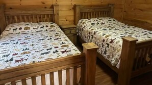 A cabin-style room with two wooden beds, each with a patterned bedspread, wooden headboards, and a small wooden nightstand between the beds. - Cozy Cabin #2 Near Lake Merrisach in Tichnor AR (Tichnor)