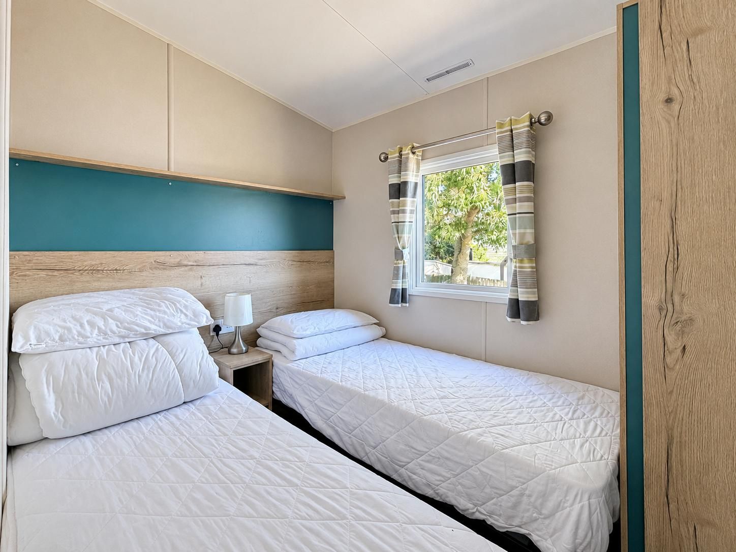 Twin bedroom with wood-effect headboards, teal accent panel, striped curtains, and a window overlooking greenery.