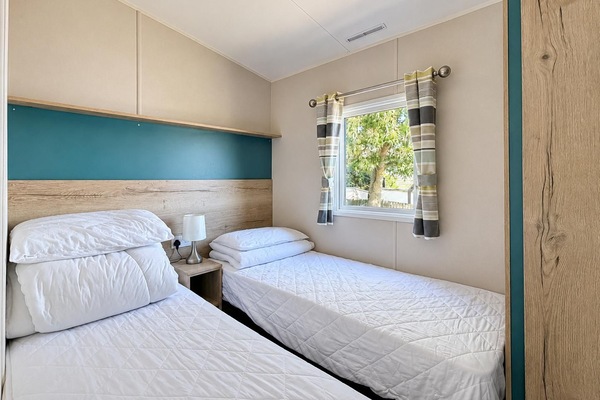 Twin bedroom with wood-effect headboards, teal accent panel, striped curtains, and a window overlooking greenery.
