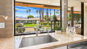 Una cocina con un amplio ventanal, un fregadero y vistas a un patio con una parrilla y palmeras.