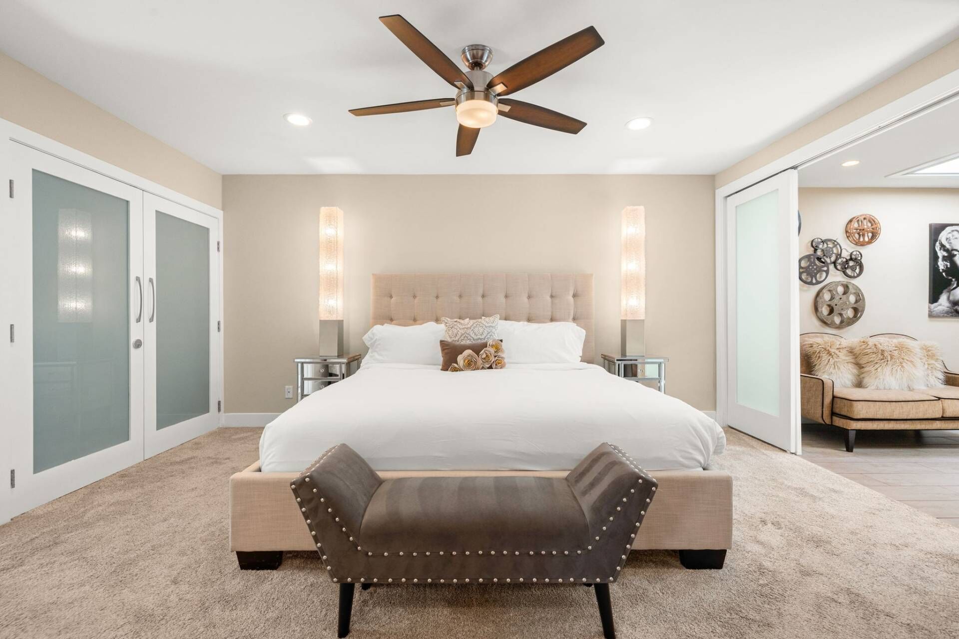 A bedroom with a bed, a ceiling fan, bedside tables, a chair, and a door with frosted glass panels.