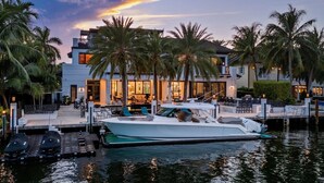 Une propriété située au bord de l’eau, avec un bateau amarré, des palmiers et une maison moderne dotée d’un espace salon extérieur.
