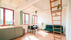 A bright room with a wooden floor, a green sofa, a dining table with chairs, and a kitchen area with a sink and cabinets. - Saint Pauls Apartment for 12 Guests (Antwerp)
