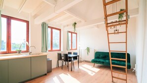 A bright room with a wooden floor, a dining table with chairs, a green sofa, a wooden ladder, and large windows with curtains. - Saint Pauls Apartment for 12 Guests (Antwerp)
