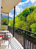 Un balcón con mesa y sillas, barandilla y vistas a árboles verdes y un paisaje distante.