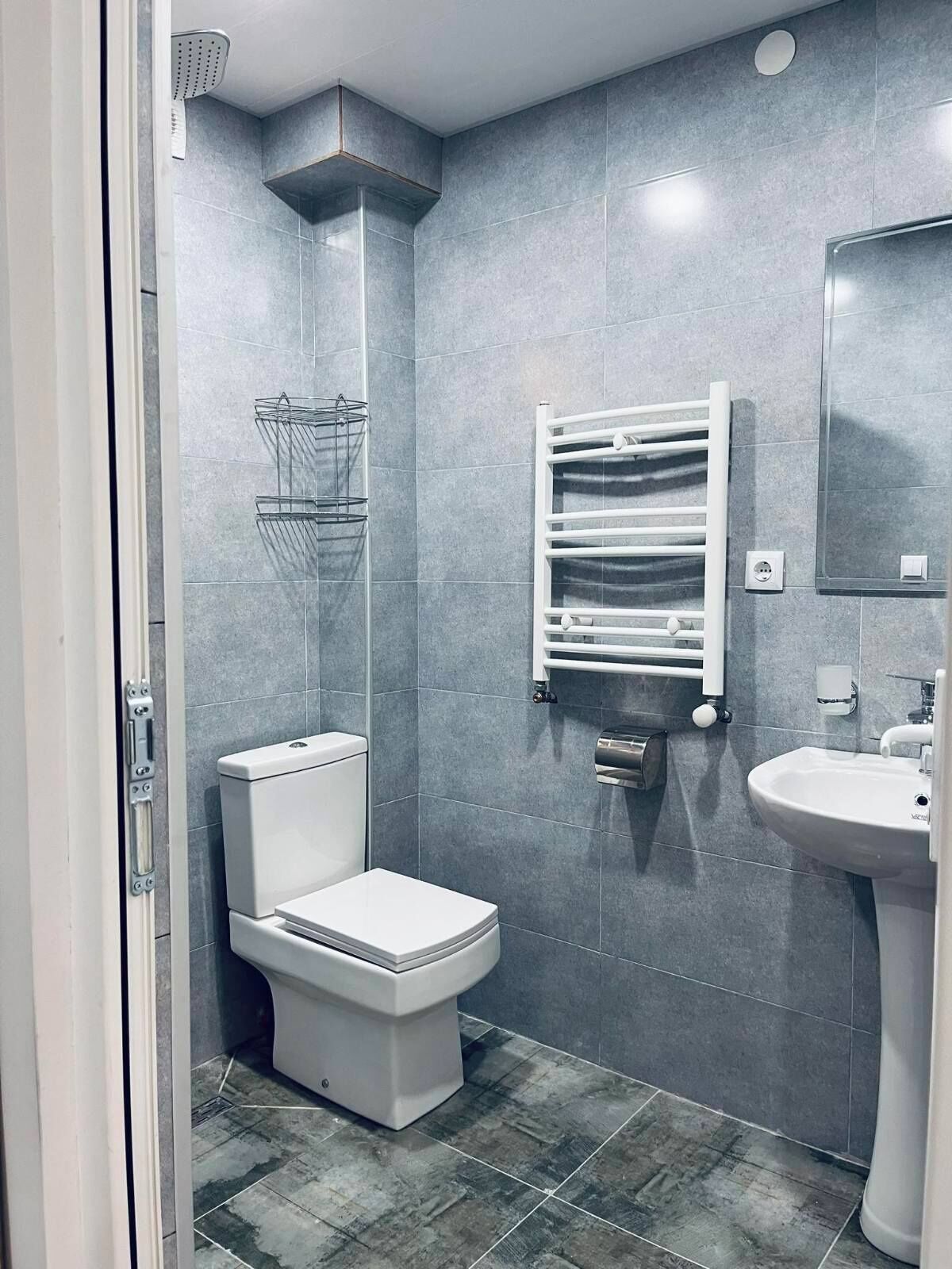 Un baño moderno con baldosas grises, un inodoro, un lavamanos, un perchero para toallas y una ducha.