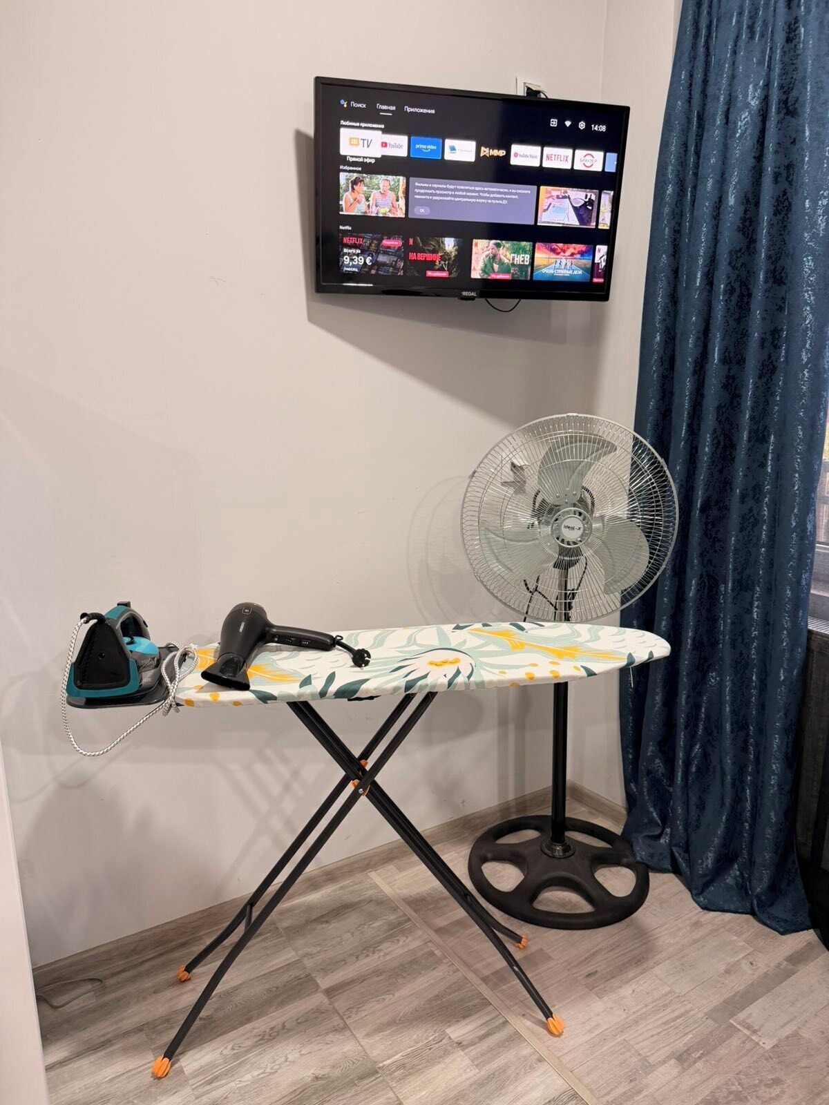 A room with a wall-mounted TV displaying streaming services, a compact ironing board with an iron, and a standing fan.